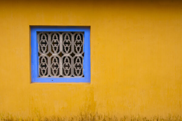 Decorative window screen with blue recessed frame, slightly off center in an orange-yellow wall.